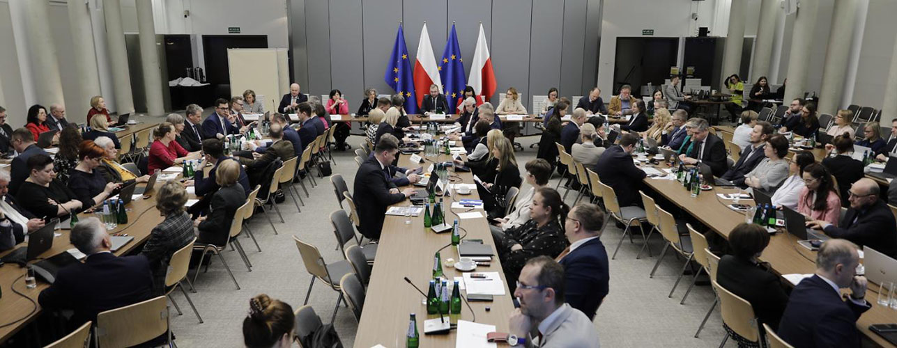 Widok sali konferencyjnej, słuchacze przy stołach ustawionych w pięciu rzędach, mówcy przy stole na tle flag UE i RP